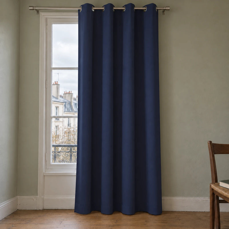 Rideau thermique uni bleu nuit à œillets, suspendu devant une fenêtre dans un intérieur aux murs gris vert.