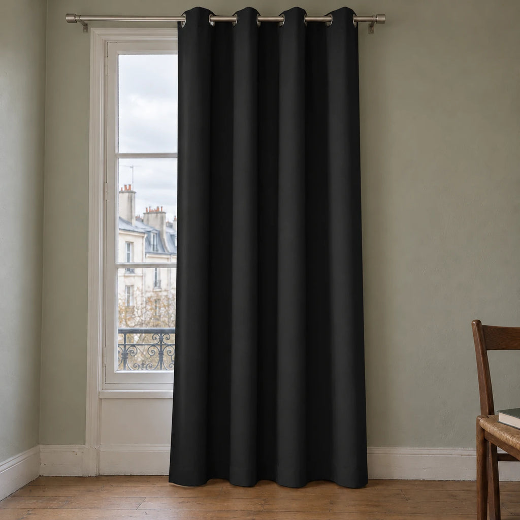 Rideau thermique uni noir à œillets, suspendu devant une fenêtre dans un intérieur aux murs gris vert.